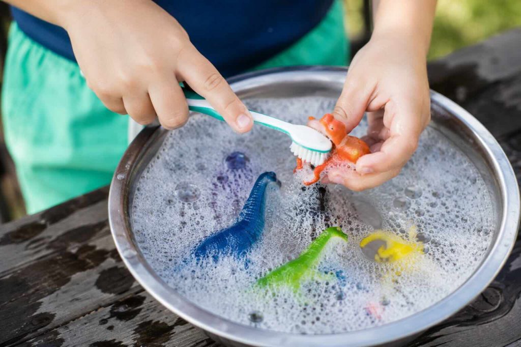 a child washing a dinosaur toy in a container of water of soap with a toothbrush